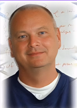 Portrait of Tony, 63, a man with a shaved head and blue eyes, wearing a blue shirt over a white undershirt, with handwritten notes in the background.
